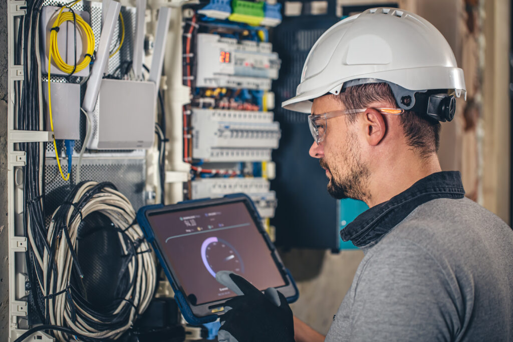 Man, an electrical technician working in a switchboard with fuses, uses a tablet.
